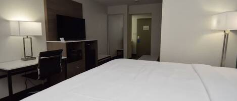 A hotel room with a large bed, a desk with a chair, a television, and a door leading to another room.