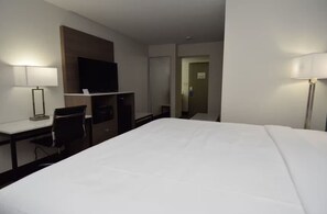 A hotel room with a large bed, a desk with a chair, a television, and a door leading to another room. - Baymont By Wyndham Batavia - Darien Lake (Elba)