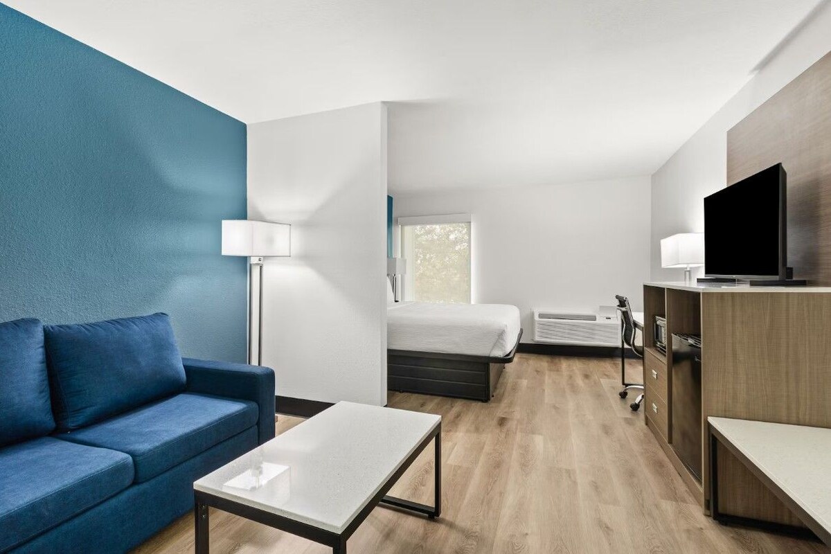 Una habitación de hotel moderna con una pared azul de acento, un sofá, una cama, un escritorio, un televisor y una ventana.