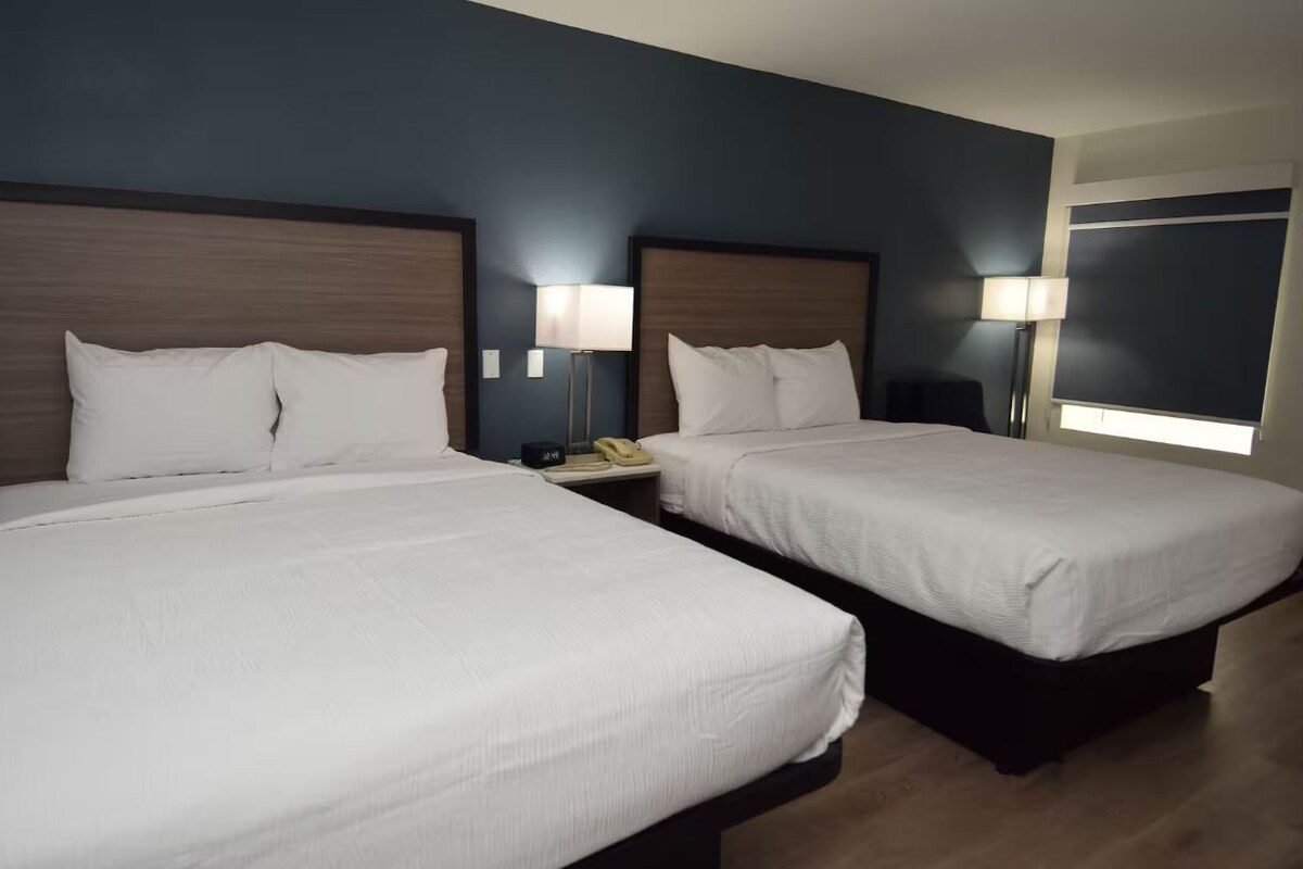A hotel room with two beds, each with a wooden headboard, white bedding, and bedside lamps.