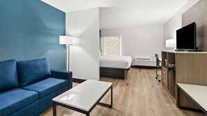A modern hotel room with a blue accent wall, a sofa, a bed, a desk, a TV, and a window. - Baymont By Wyndham Batavia - Darien Lake (Elba)