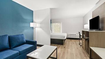 A modern hotel room with a blue accent wall, a sofa, a bed, a desk, a TV, and a window.
