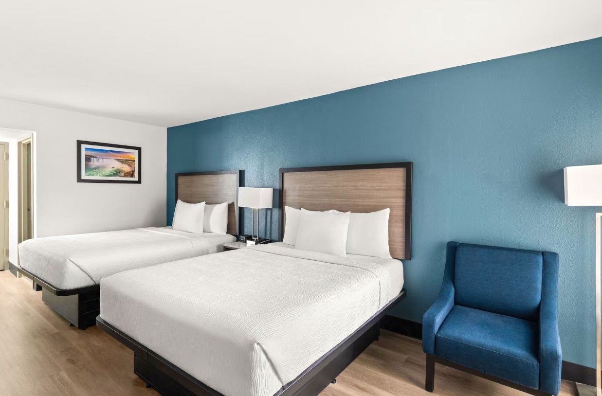 A hotel room with two beds, a blue armchair, and a framed picture on the wall.