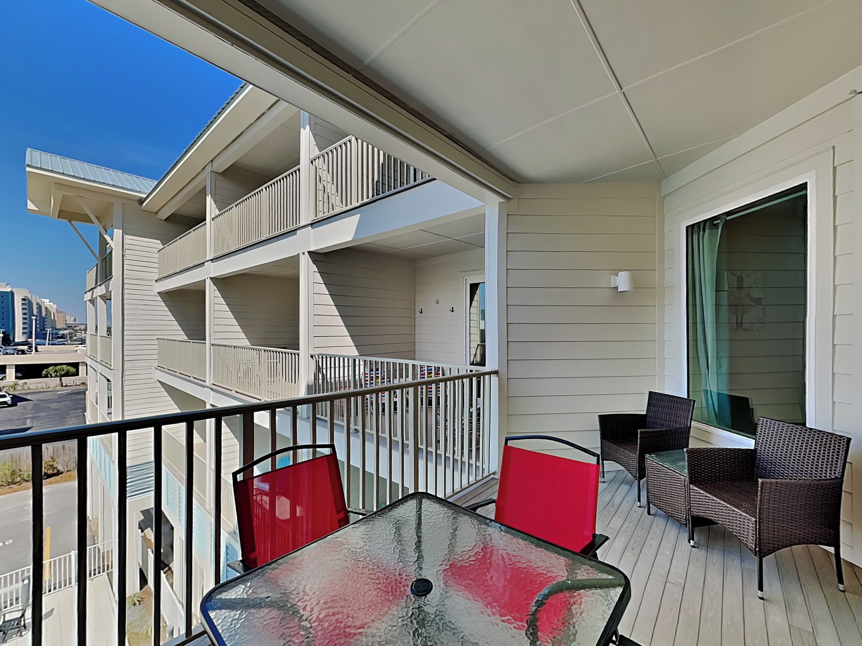 A balcony with a glass table, red chairs, wicker chairs, and a view of a cityscape.