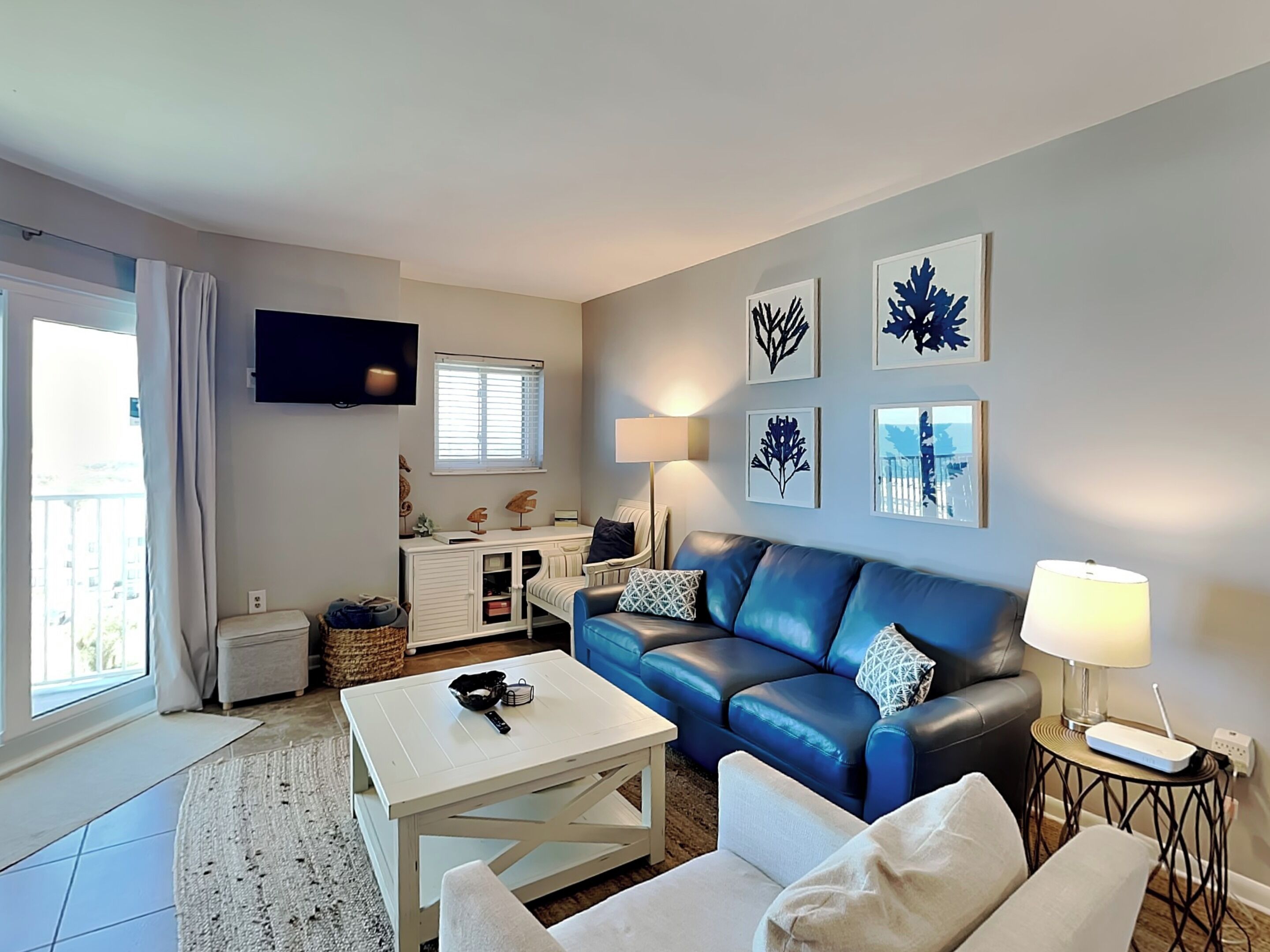 Ein modernes Wohnzimmer mit einem blauen Ledersofa, einem weißen Couchtisch und einem an der Wand montierten Fernseher. An den Wänden befinden sich Bilder in Rahmen und ein Fenster mit Jalousien.