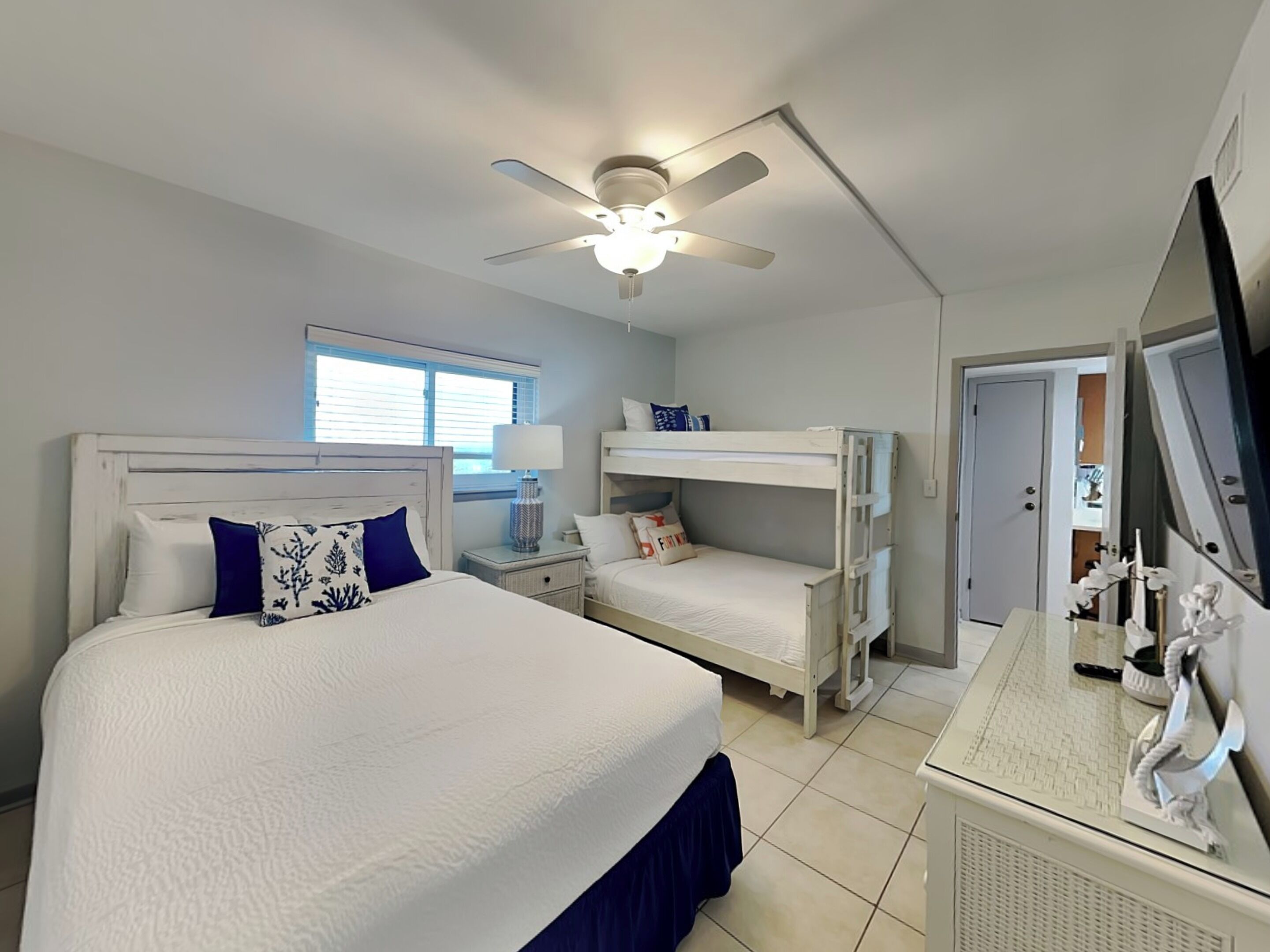 A bedroom with a large bed, a smaller bunk bed, a ceiling fan, a window, and a dresser with a mirror.