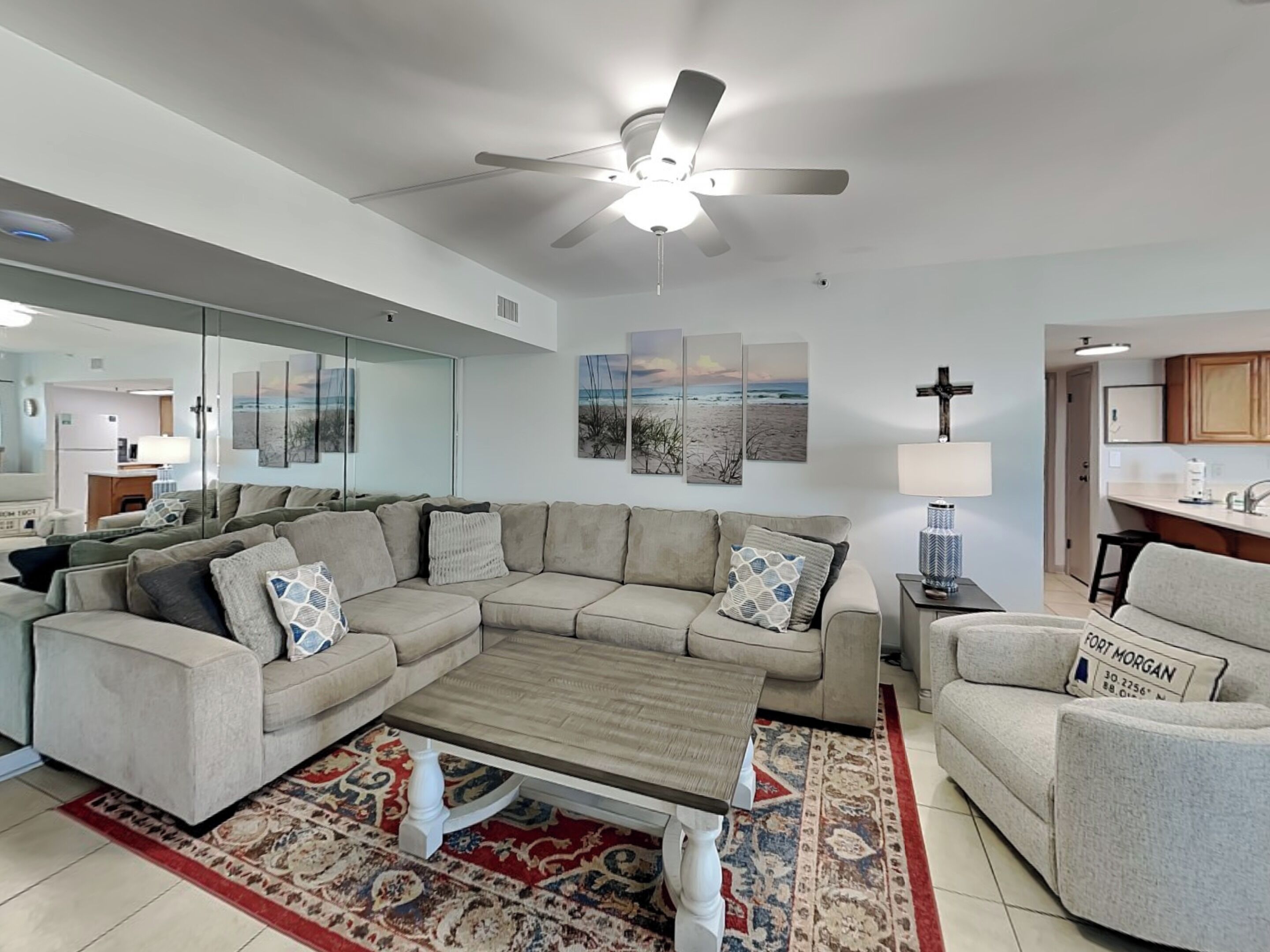 A modern living room with a sectional sofa, a coffee table, and a patterned rug. There is a ceiling fan, a kitchen area in the background, and a cross on the wall.