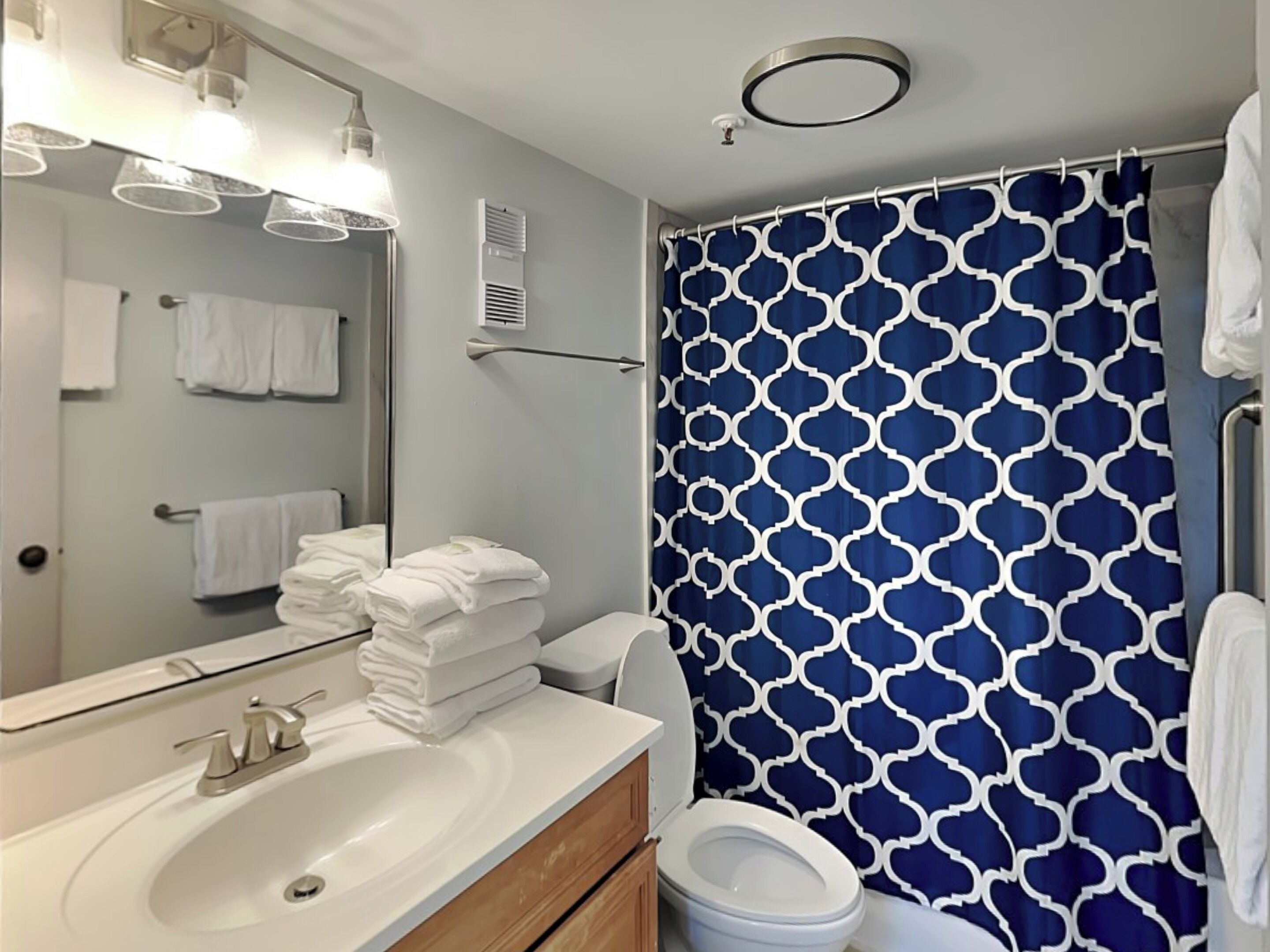 Un baño con lavamanos blanco, un espejo, inodoro y cortina de ducha azul estampada.