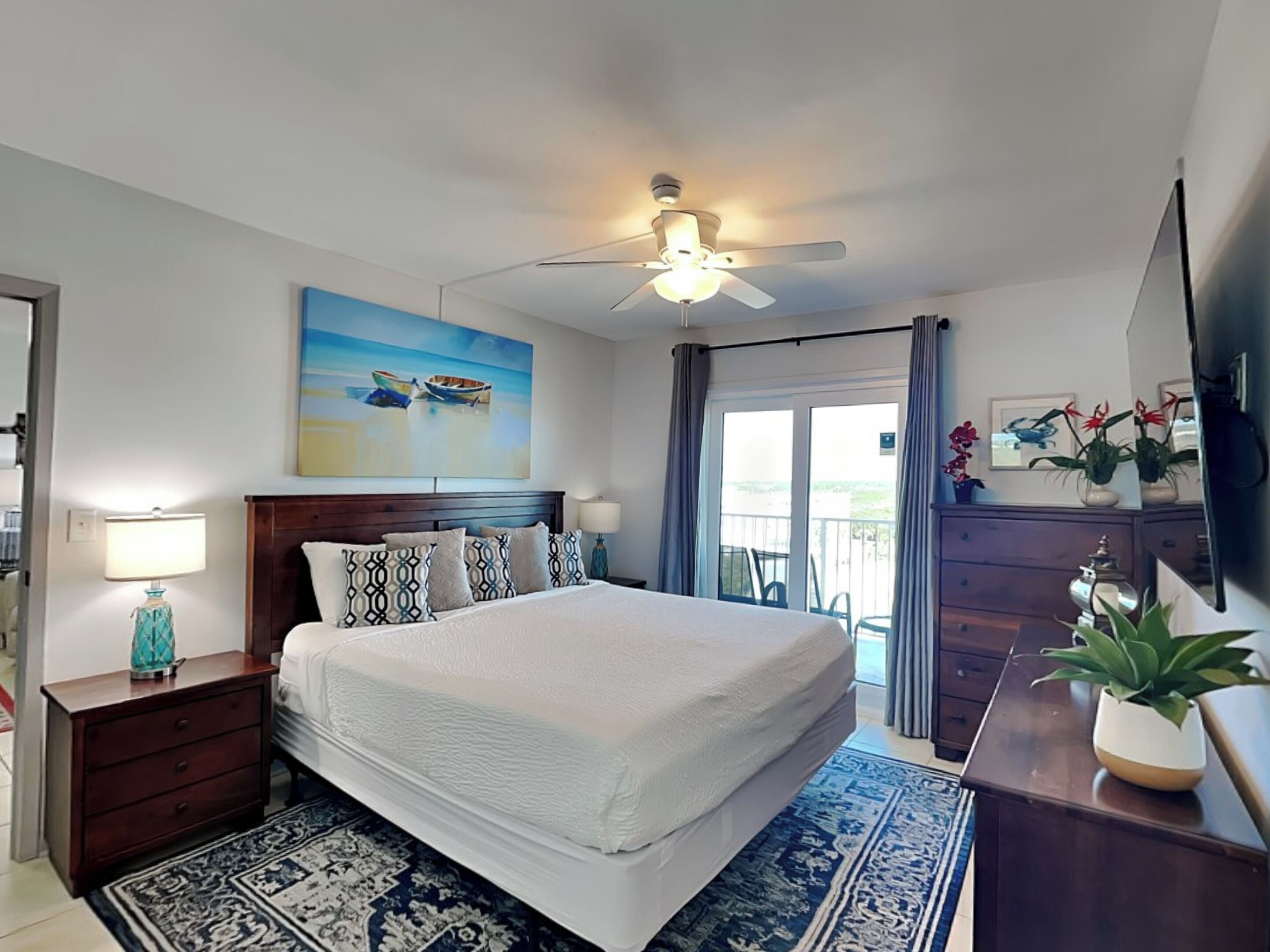 Une chambre d’hôtel avec un grand lit, un ventilateur de plafond, un tableau représentant des bateaux, un balcon avec vue et un téléviseur fixé au mur.