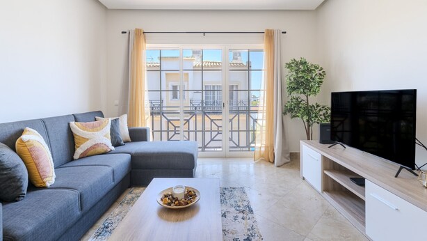 Una sala de estar moderna con un sofá gris, una mesa de centro con un cuenco de fruta, un televisor de pantalla plana sobre un mueble de madera y un balcón con vista a edificios.