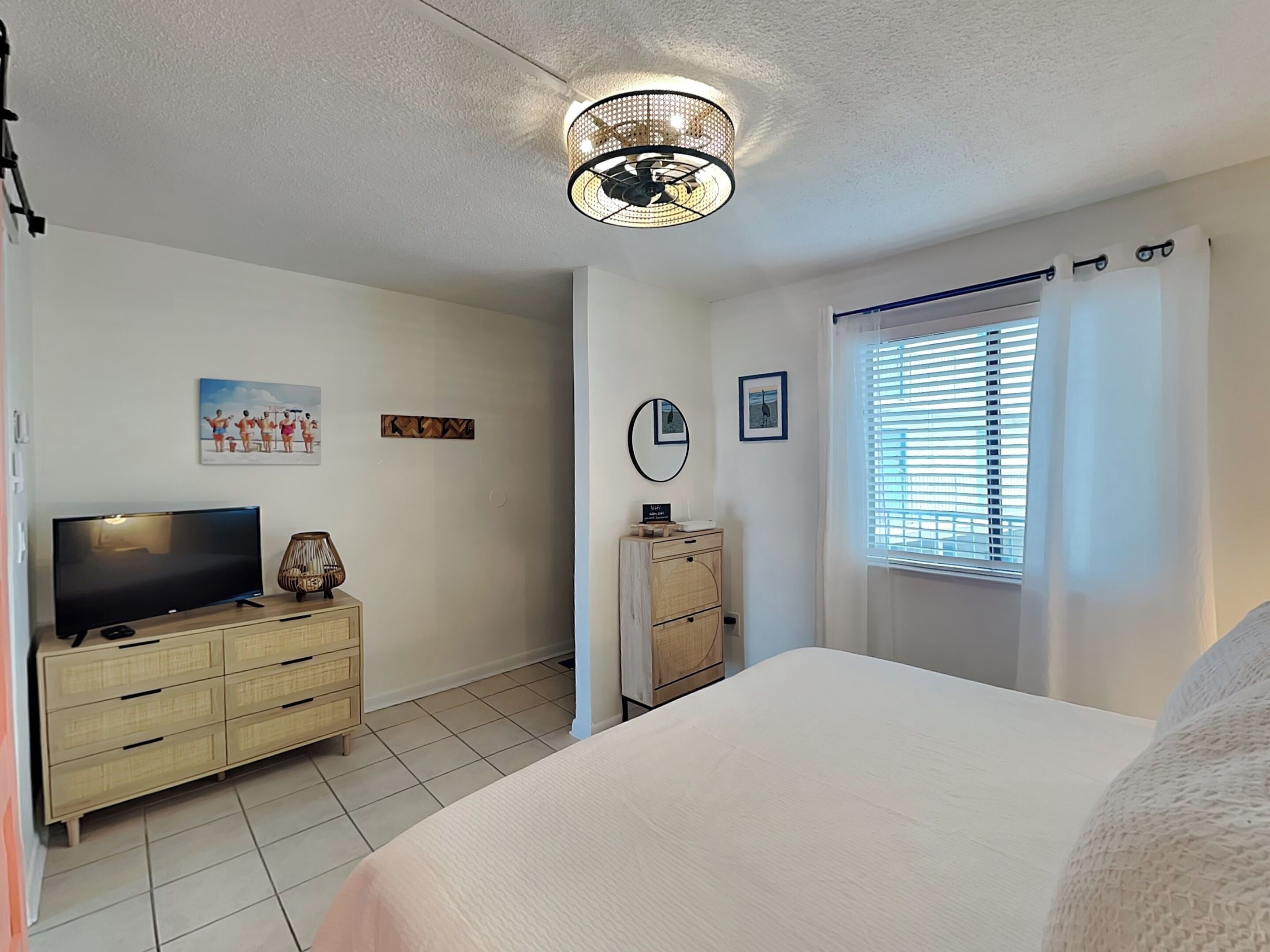 A bedroom with a bed, a TV on a wooden dresser, a ceiling light fixture, and a window with blinds.