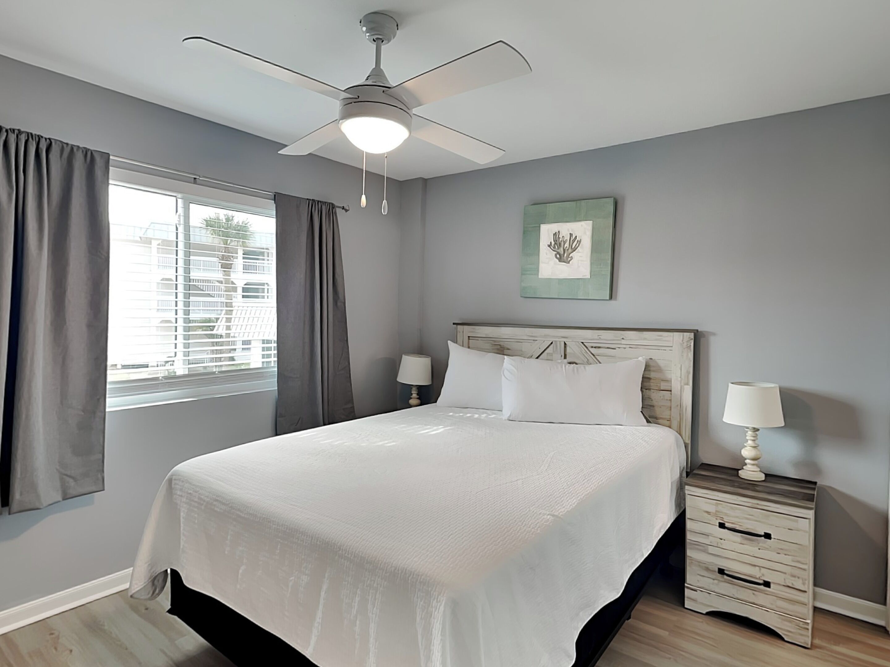 A bedroom with a bed, a ceiling fan, a nightstand with a lamp, a window with curtains, and a framed picture on the wall.