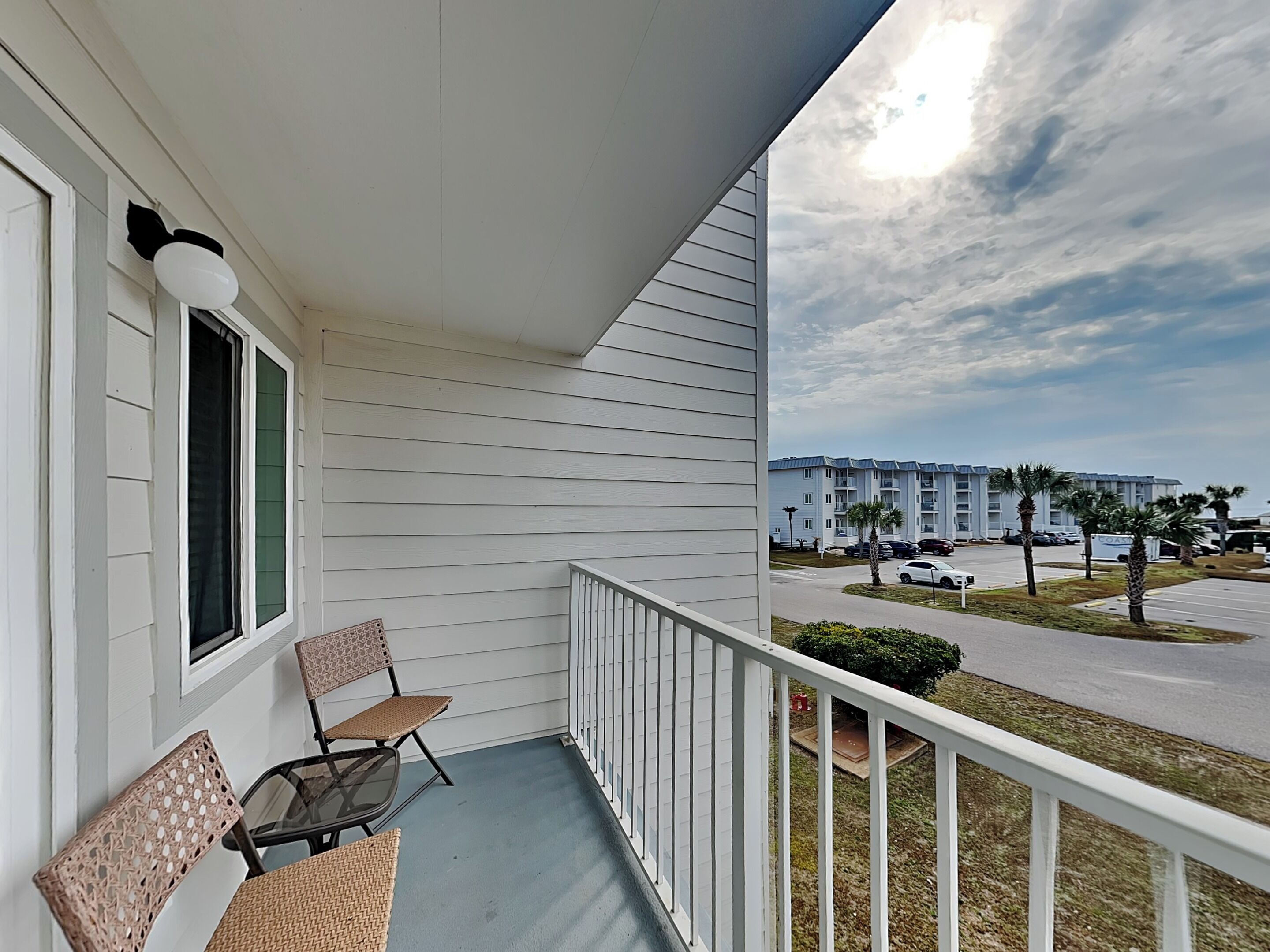 A balcony with wicker furniture, a white railing, and a view of a parking lot and buildings in the background.