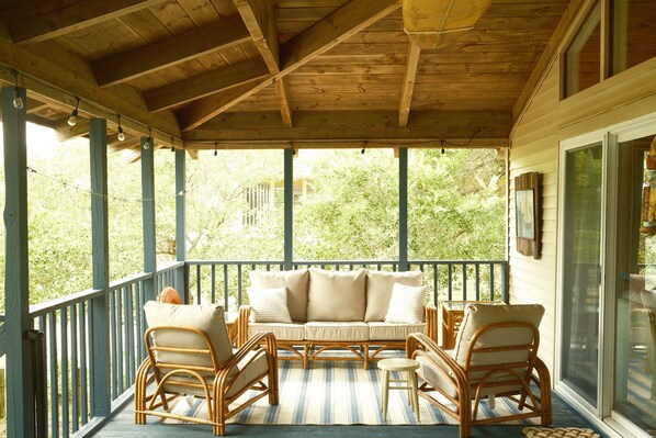 A covered porch with a sofa and wicker chairs, wooden beams, and a view of greenery. - Nautical by Nature | Restorative Coastal Retreat w/ Hot Tub & Cold Plunge (Kill Devil Hills)