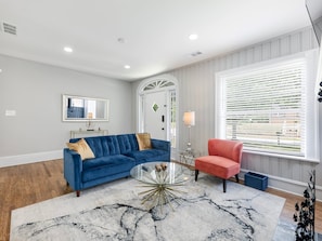 Una sala de estar con un sofá azul, un sillón rojo, una mesa de centro de vidrio y un ventanal con persianas.