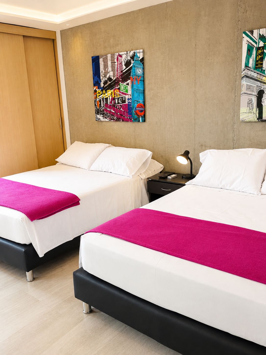 Une chambre d’hôtel avec deux lits, chacun agrémenté de draps blancs et d’un plaid rose. Un tableau représentant un paysage urbain est accroché au mur, et une table de chevet avec une lampe se trouve à côté du lit.