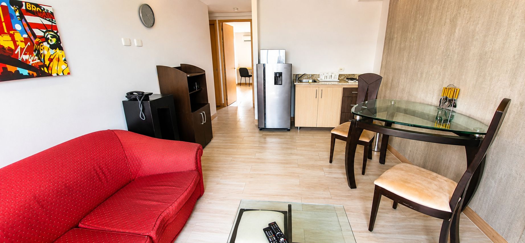 Un espacio habitable compacto con un sofá rojo, una mesa de comedor de cristal y una zona de cocina con refrigerador y armarios.