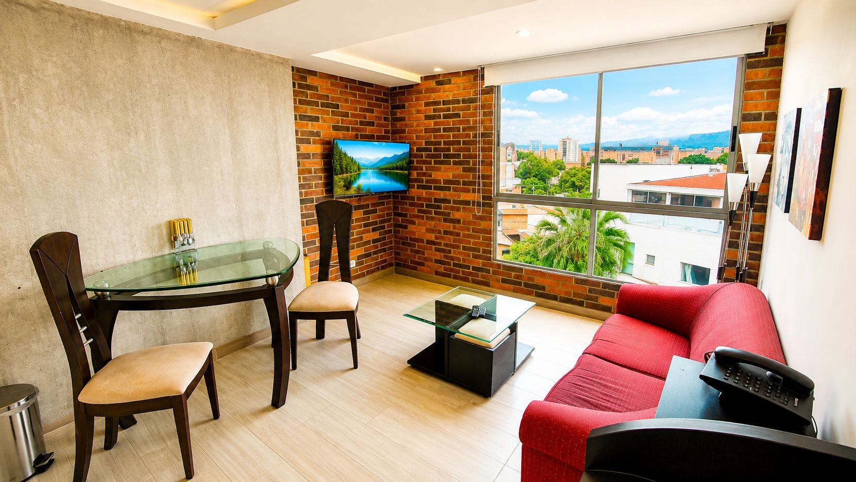 A modern living room with a glass table, red sofa, and a TV mounted on a brick wall.