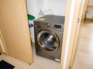 A compact, stainless steel front-loading washing machine in a small laundry room with a wooden door and a white wall.