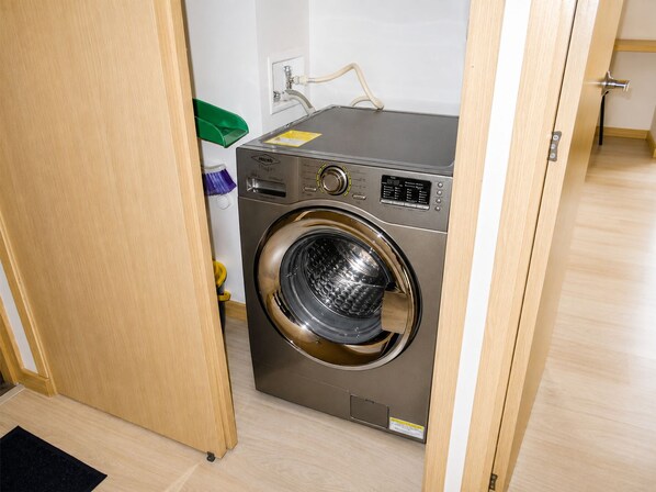 Lavadora de carga frontal, compacta y de acero inoxidable, ubicada en un lavadero pequeño con puerta de madera y pared blanca.