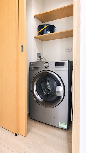 Máquina de lavar compacta e moderna, com painel de controle, em uma pequena lavanderia com prateleiras de madeira acima.