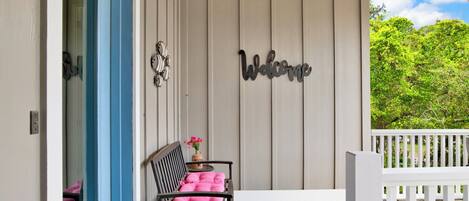 A wooden deck with a white railing, a black chair with a pink cushion, and a 'Welcome' sign on the wall.