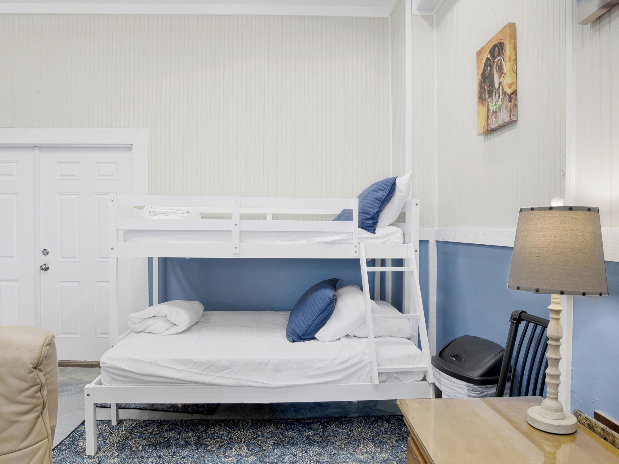 A bunk bed with white bedding, a blue pillow, and a small table with a lamp.