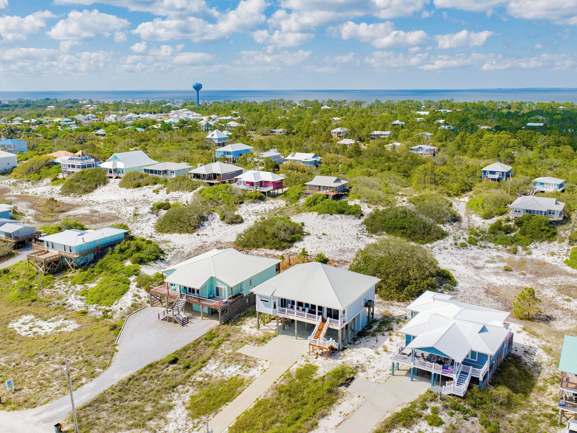 An aerial view of a coastal residential area with houses on stilts, a water tower, and a clear view of the ocean.