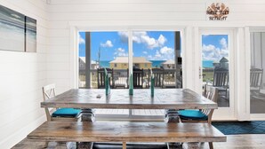 Un coin repas avec une grande table et des chaises en bois, une vue sur la plage et l’océan par de grandes fenêtres, et une enseigne indiquant « The Bare Feet Retreat ».