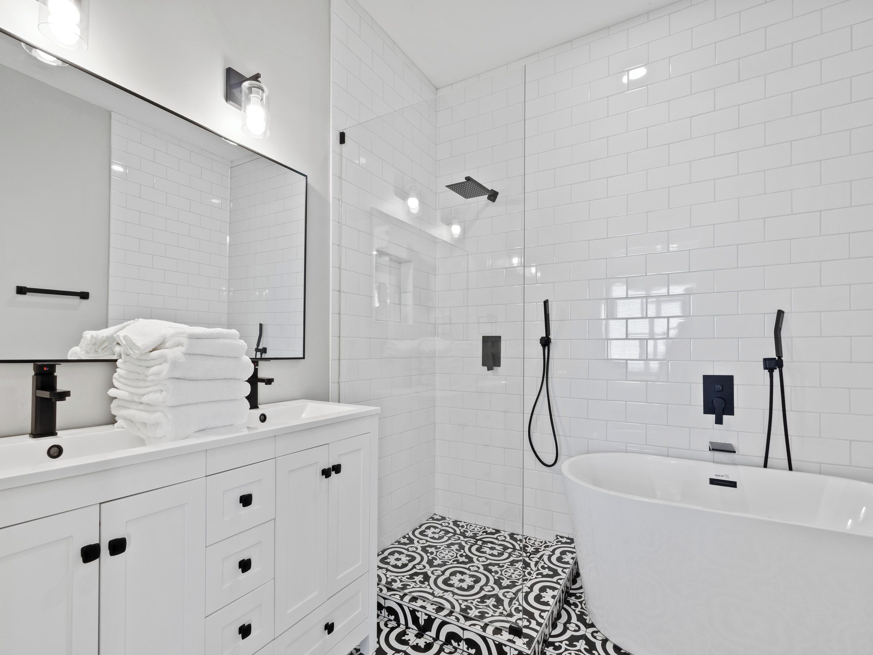 A modern bathroom with white tiles, a large mirror, a sink with a towel stack, a shower with a handheld showerhead, and a bathtub.