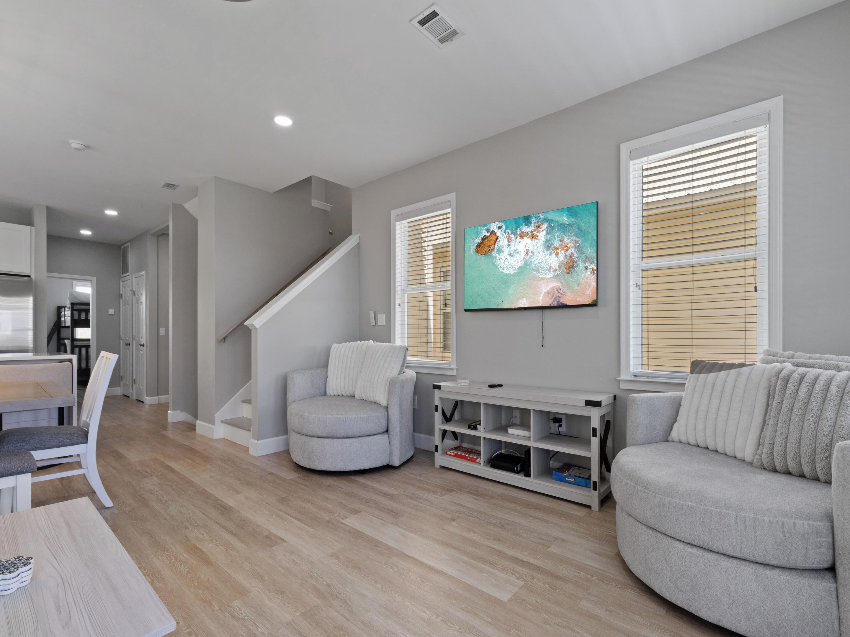 A modern living room with a grey sofa, a white armchair, a flat-screen TV mounted on the wall, and a staircase leading to another level.