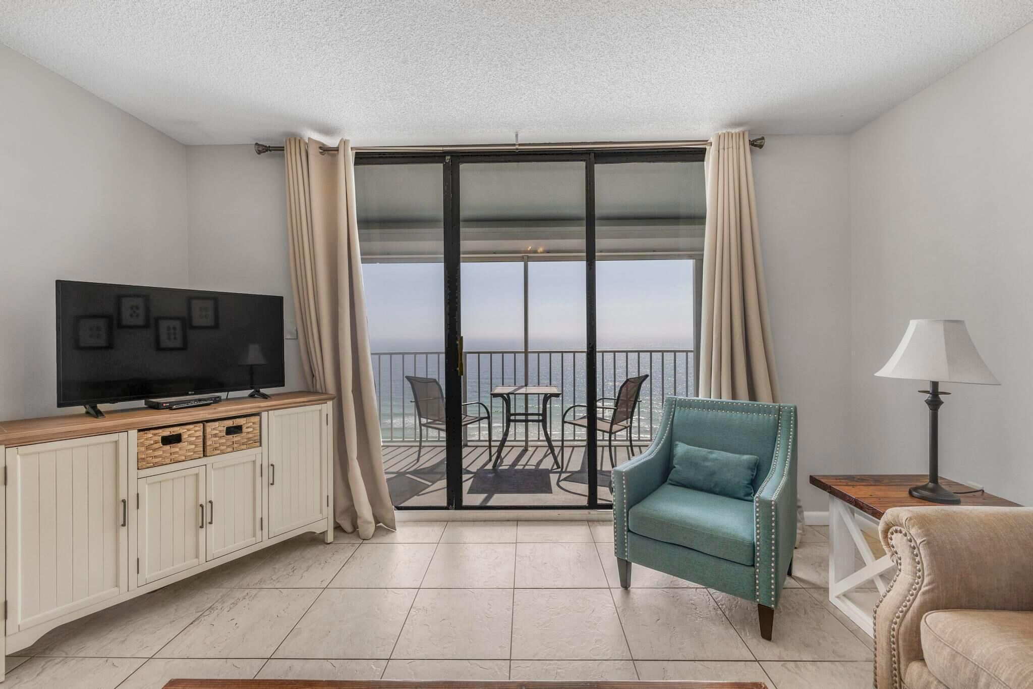 A hotel room with a TV on a wooden cabinet, a teal armchair, a small table with a lamp, and a balcony with a table and chairs.