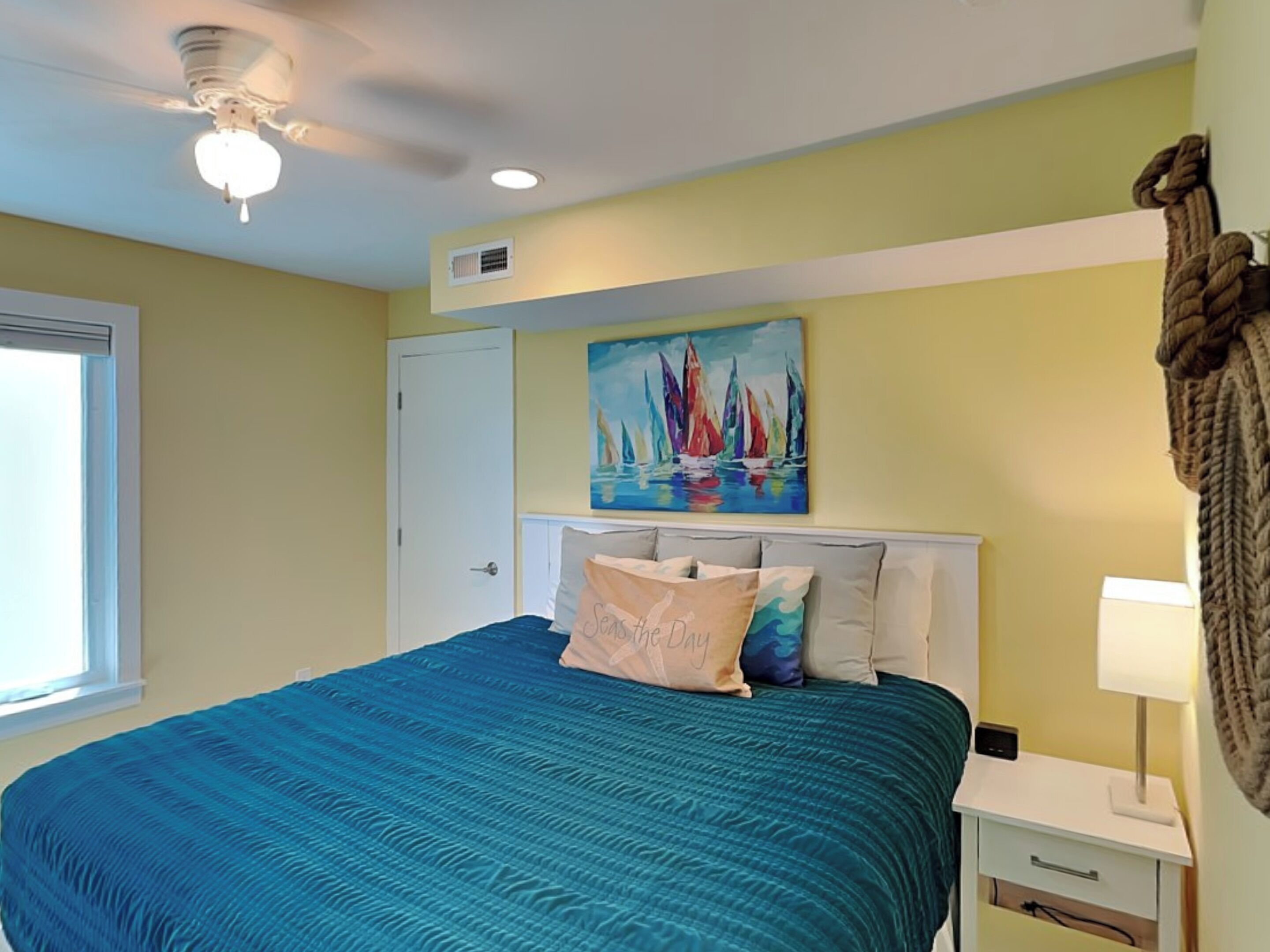Une chambre à coucher avec un lit, un ventilateur de plafond, un tableau représentant des voiliers, une fenêtre et une table de chevet avec une lampe.