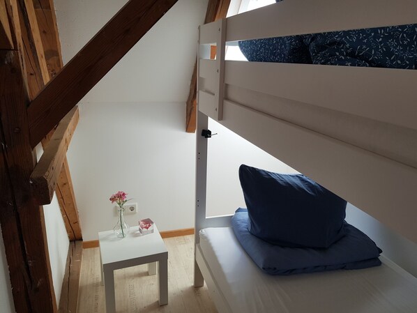 Une chambre de type dortoir avec des lits superposés, une petite table blanche ornée d’un vase et une fenêtre donnant sur le ciel.