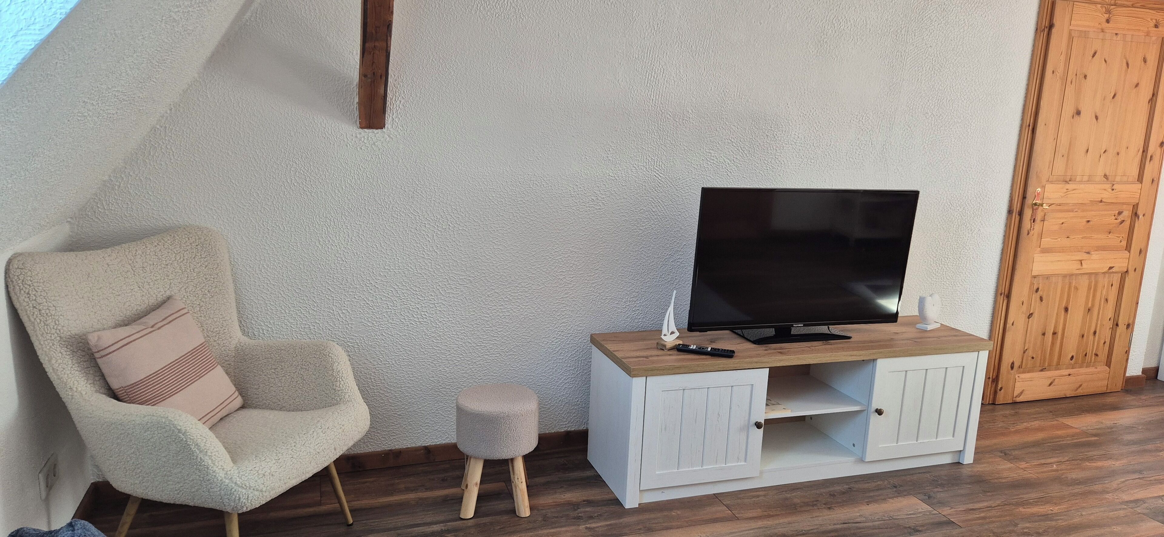 Ein gemütliches Zimmer mit bequemem Sessel, kleinem Hocker und einem Holz-Fernsehschrank mit Flachbildfernseher.
