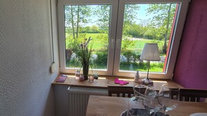 Ein Zimmer mit großem Fenster und Blick auf eine grüne Landschaft, einem Heizkörper, einem Holztisch mit Lampe sowie einer Vase mit Blumen.