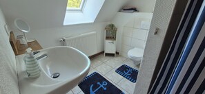 Une petite salle de bain avec un lavabo blanc, un miroir, un radiateur, des toilettes et une petite fenêtre.
