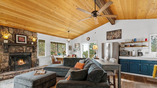 A cozy living room with a fireplace, a large sectional sofa, and a kitchen area with blue cabinets and a wooden ceiling.