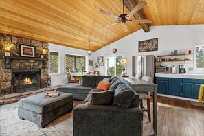 A cozy living room with a fireplace, a large sectional sofa, and a kitchen area with blue cabinets and a wooden ceiling. - Top-Rated Treehouse Retreat with Hot Tub & Mountain Views, Pet Friendly (Big Bear)
