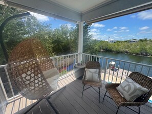A covered outdoor deck with wicker furniture and a swing chair, overlooking a body of water and greenery. - Cozy 2-bedroom house in charming Big Pine Key with canal view and relaxing pool (Big Pine Key)