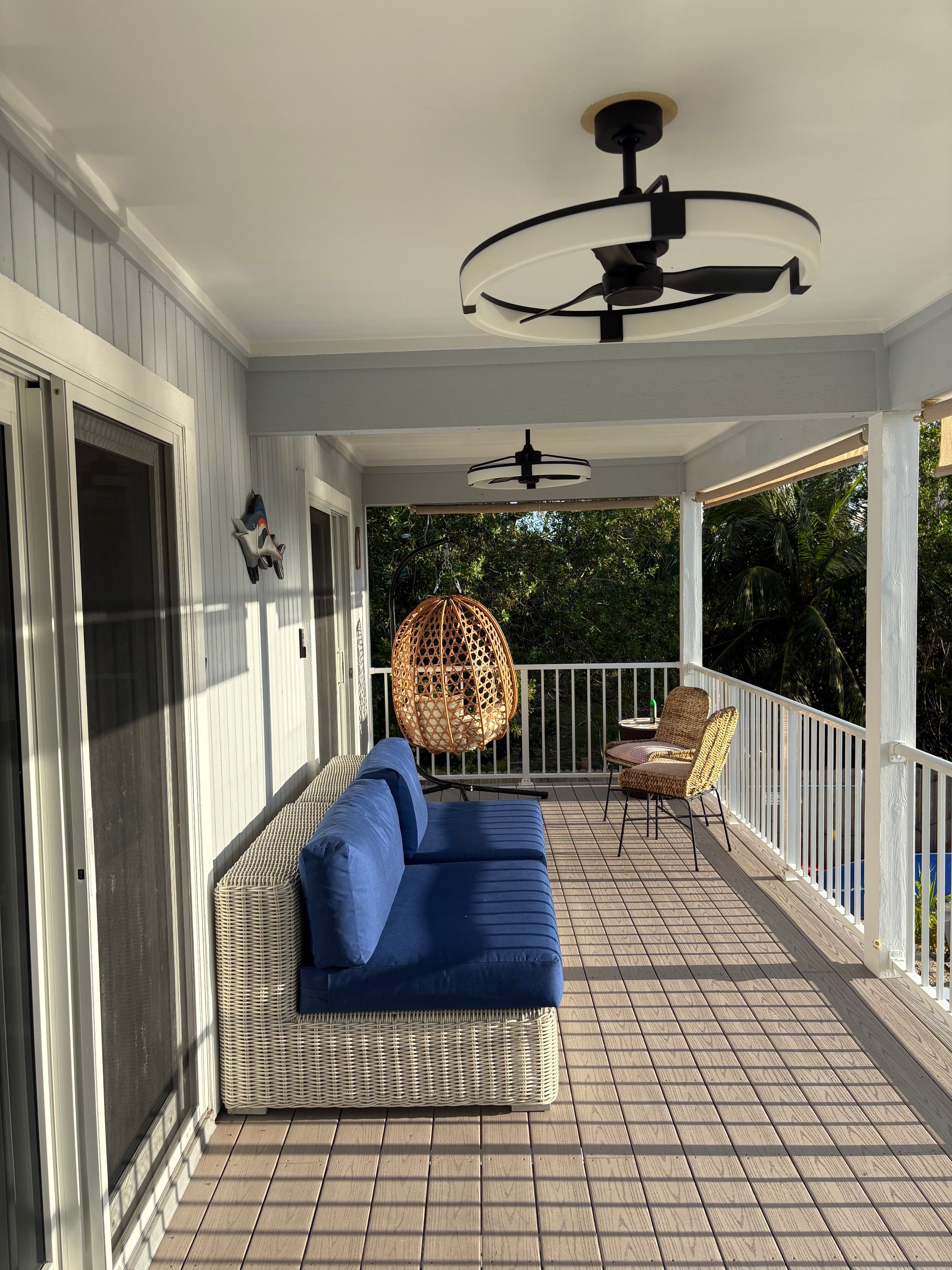Ein überdachter Außenbereich mit Deckenventilator, einem Korbsessel mit blauen Kissen und einem hängenden Rattan-Sessel.