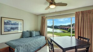 Une chambre à coucher avec un lit, une table et une chaise. Il y a une fenêtre avec des rideaux, et de là, on peut voir un lac et des maisons.