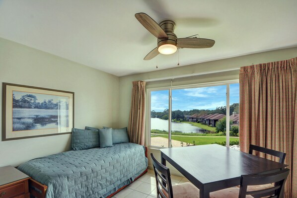 A bedroom with a bed, a table, and a chair. There is a window with curtains, a ceiling fan, and a framed picture on the wall.