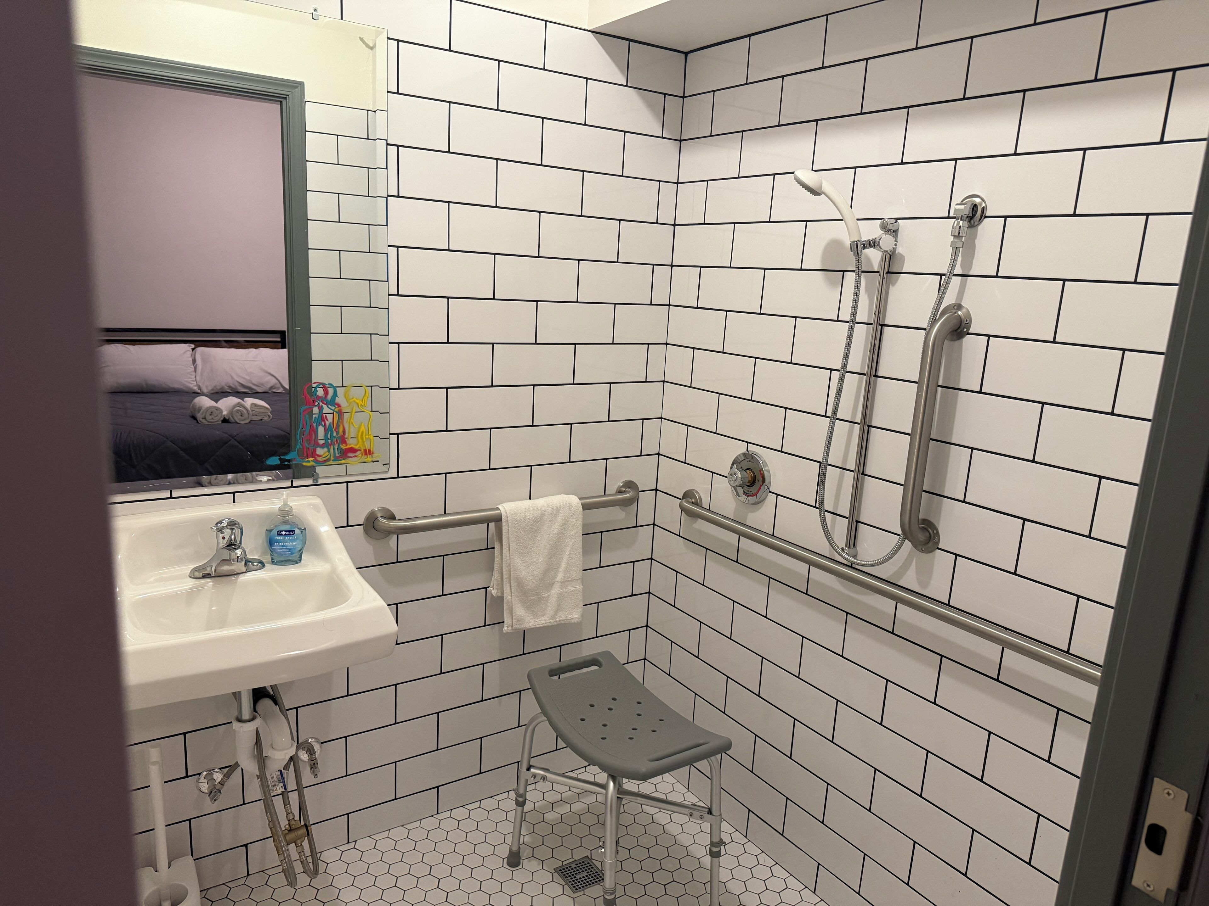 A bathroom with white subway tiles, a sink, a mirror, a shower with a handheld showerhead, and a grab bar.
