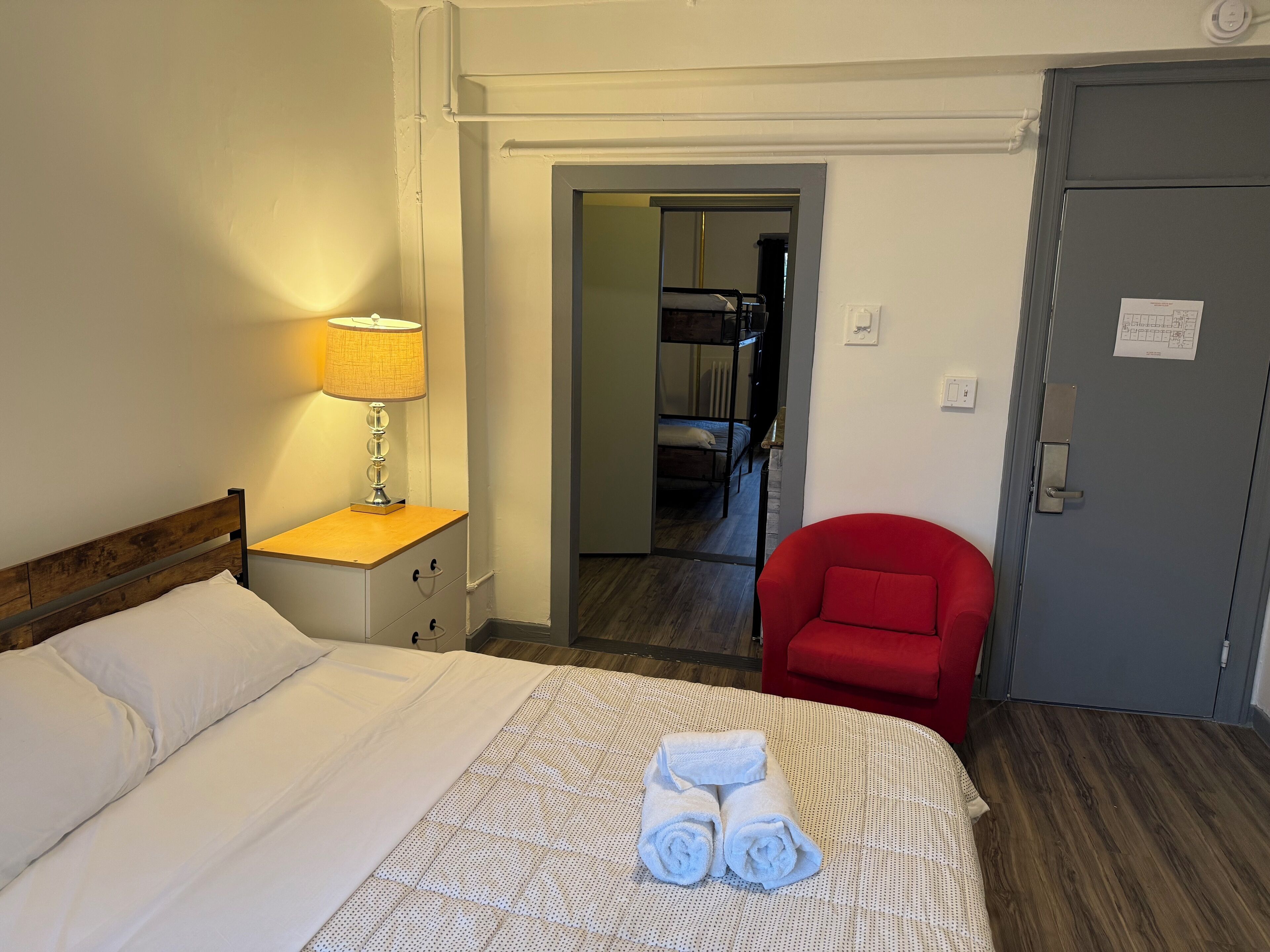 A hotel room with a bed, a red chair, a lamp, and a door.
