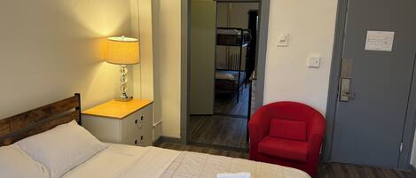 A hotel room with a bed, a red chair, a lamp, and a door.