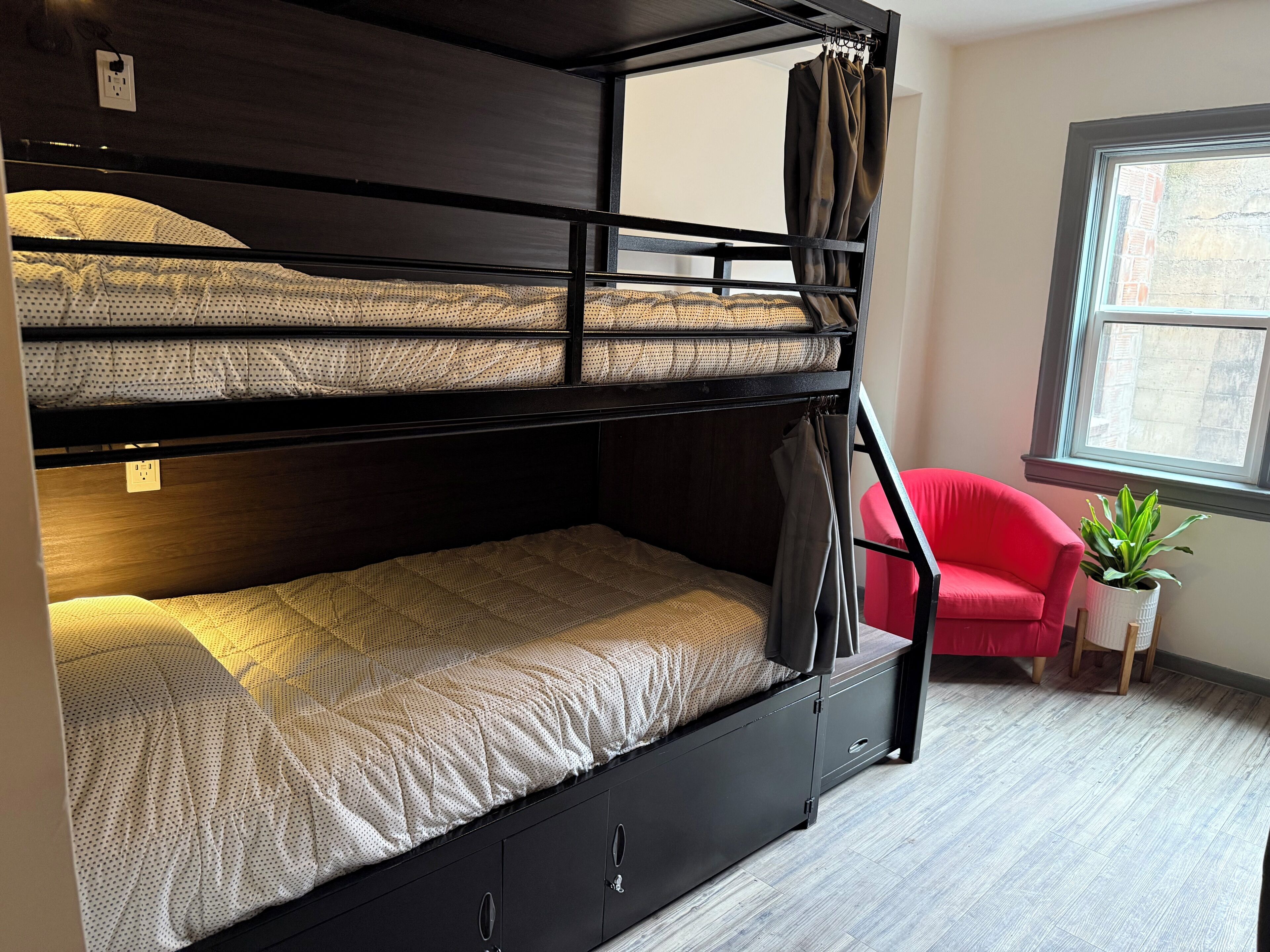 Un lit superposé avec un lit inférieur et un bureau escamotable, une chaise rouge et une plante en pot près d’une fenêtre.