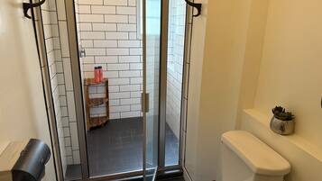 A modern bathroom with a glass shower enclosure, white tiled walls, a wooden shelf with toiletries, and a toilet with a decorative plant.