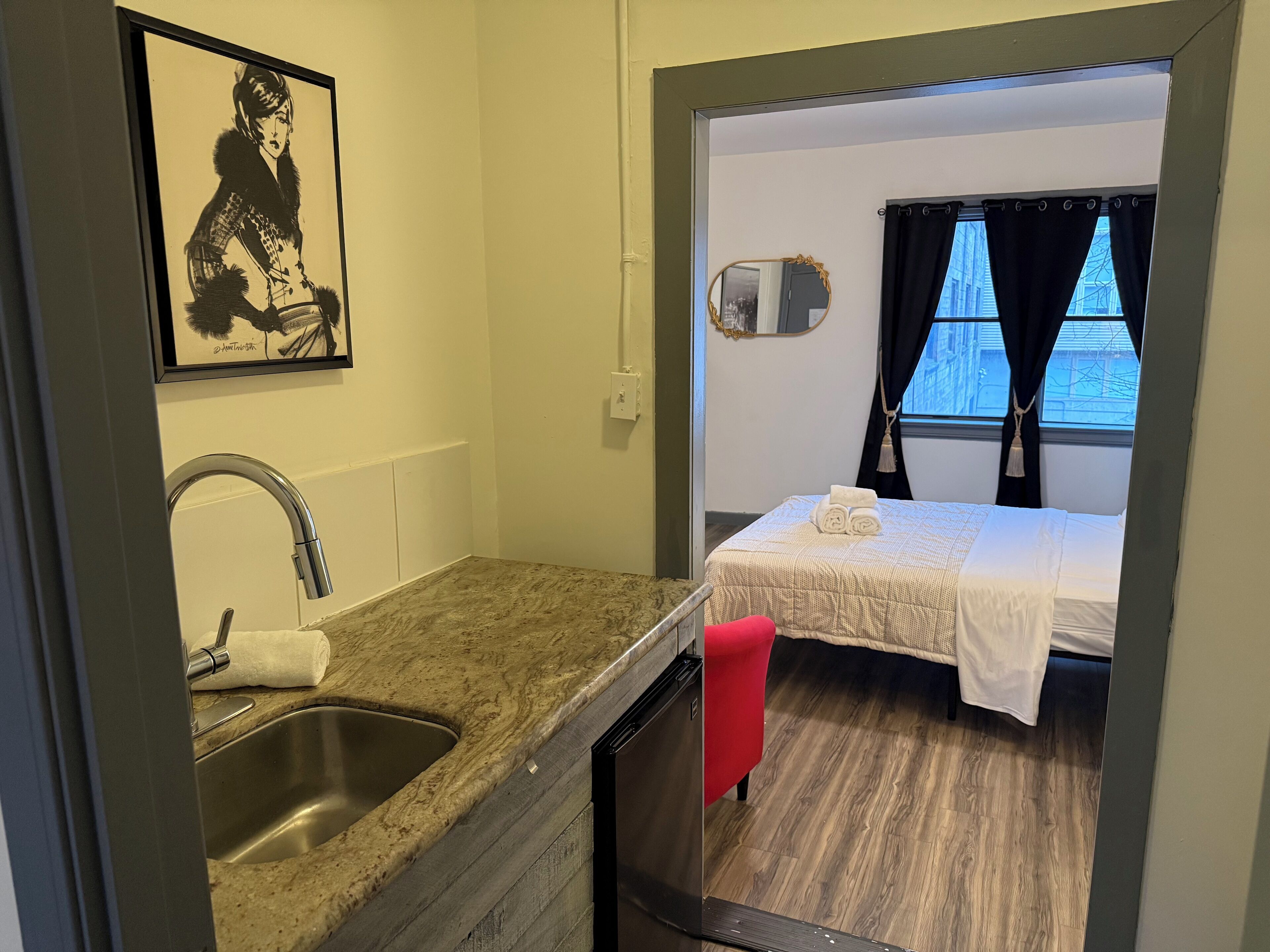 A compact room with a kitchenette, a bed with a white bedspread, a window with curtains, and a framed artwork on the wall.