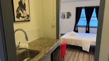A compact room with a kitchenette, a bed with a white bedspread, a window with curtains, and a framed artwork on the wall.